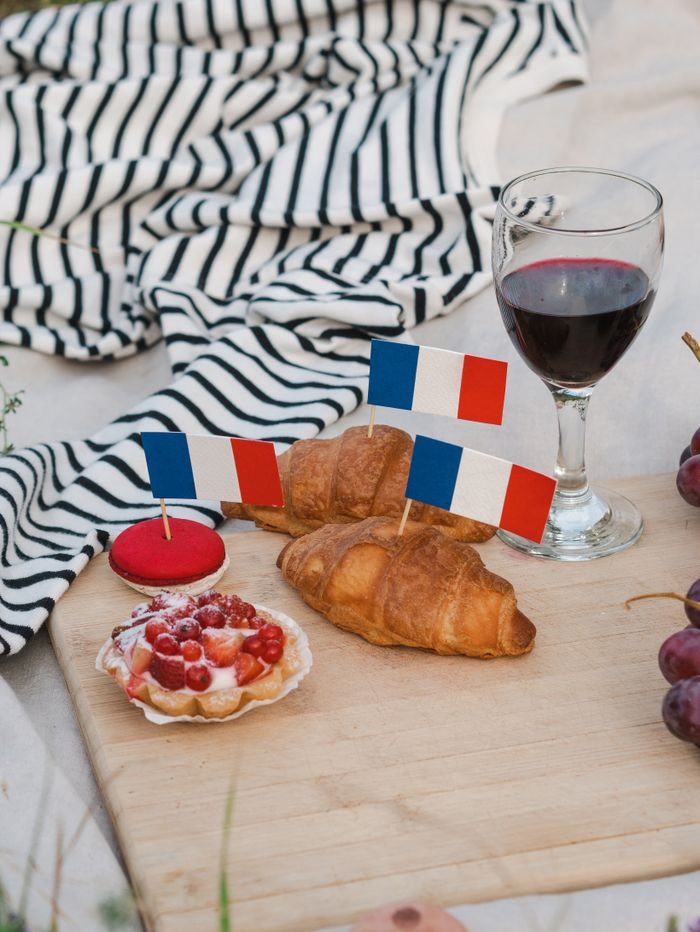 French-themed picnic setup with croissants, wine, and miniature French flags on a wooden board.
